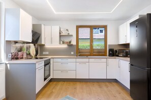 Eine moderne Küche mit weißen Schränken, einem eingebauten Ofen, einem Kühlschrank und einem Fenster mit Blick auf ein Haus.