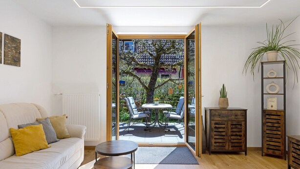 A modern living room with a sofa, a small round table, and a view of an outdoor patio with a tree and a building in the background. - Apartment “Am Brändbach” with Private Terrace, Garden, and Wi-Fi (Bräunlingen)