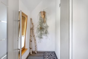 Ein schmaler Flur mit einer Holztür, einem Fenster mit Rollläden, einer gemusterten Matte und einer hängenden Pflanze.