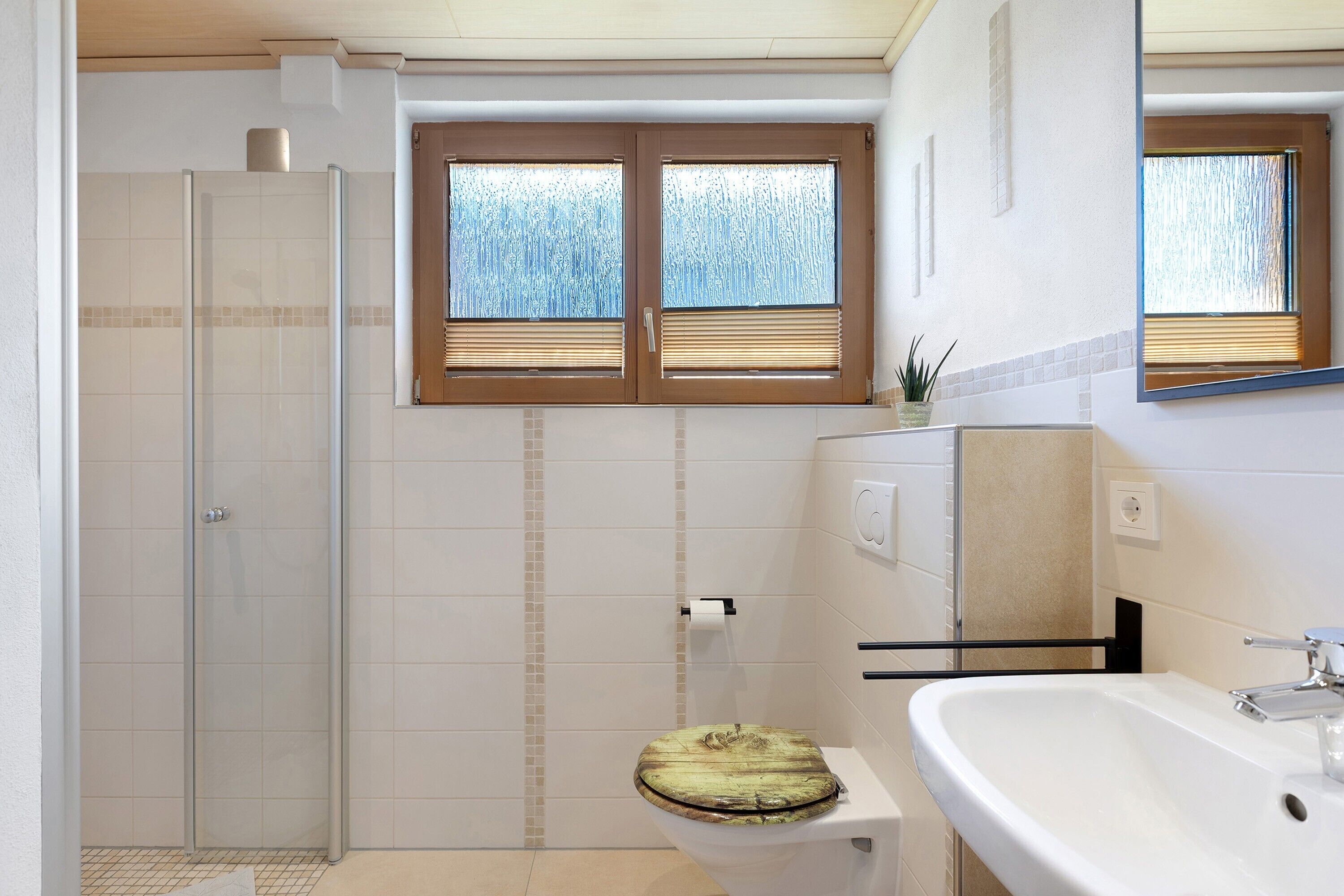 Une salle de bain avec une douche à parois de verre, des toilettes avec un abattant en bois, un lavabo et deux fenêtres équipées de stores.