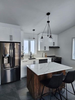 A modern kitchen with a stainless steel refrigerator, a central island with bar stools, and pendant lighting. - Chalet La Félicita, enchanting waterfront. (Saint-Adolphe-d'Howard)