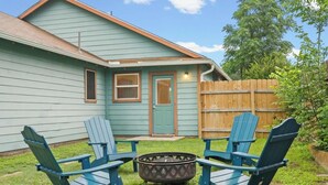 Een huis met een voordeur, twee ramen, een houten hek en twee blauwe Adirondack-stoelen rond een vuurkorf.
