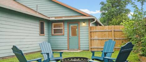 A house with a front door, two windows, a wooden fence, and two blue Adirondack chairs around a fire pit.