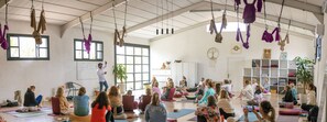 Un cours de yoga dans une pièce spacieuse, dotée d’un haut plafond, de grandes fenêtres et de tapis de yoga suspendus.
