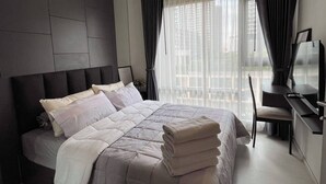 A modern bedroom with a large bed, a desk, and a view of the city through sheer curtains. - Luxury Condo in Khlong Toei City Views & Skytrain Access (Bangkok)