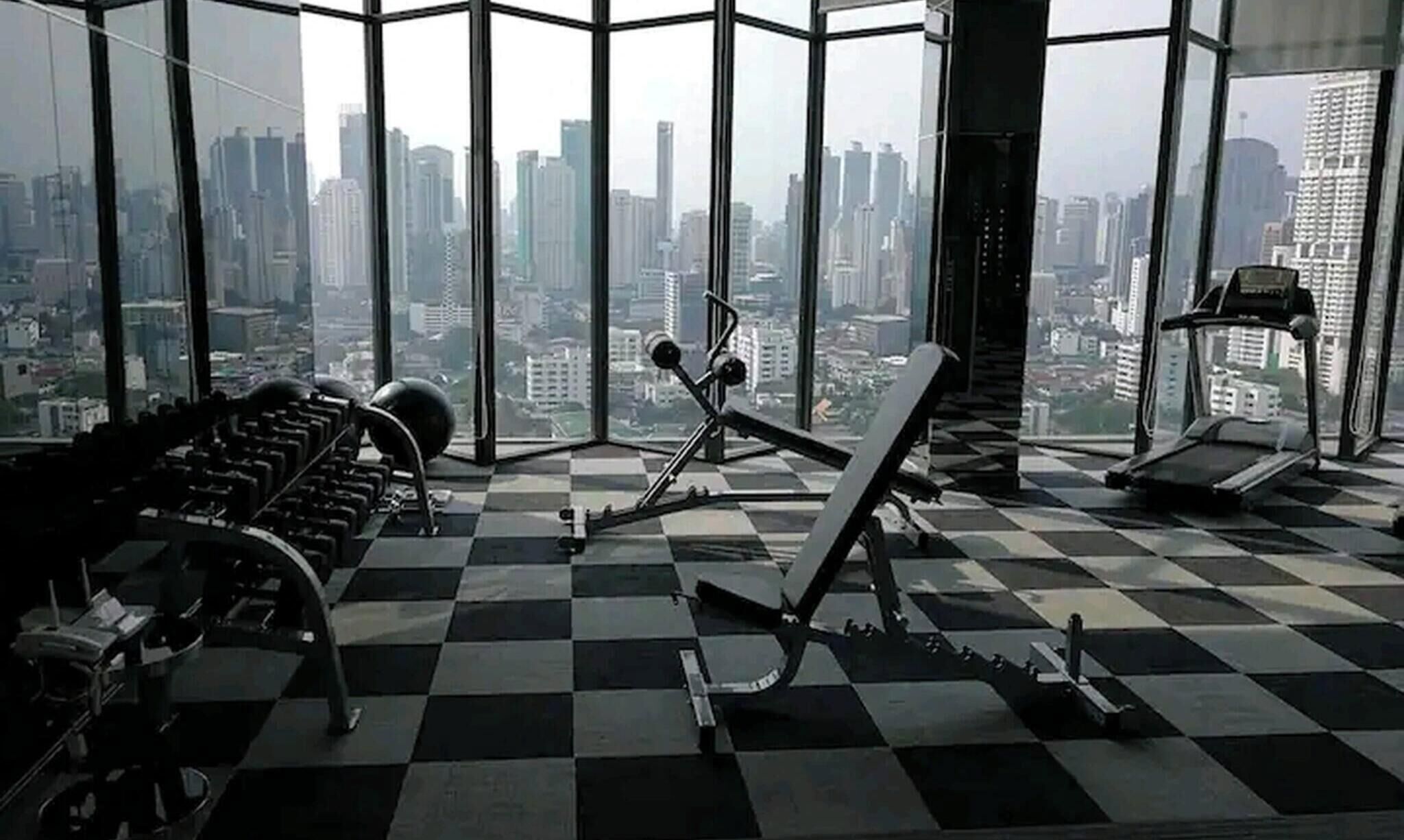 A gym with a checkered floor, exercise equipment, and a city view through large windows.