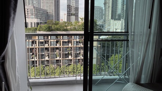A high-rise building with a view of other tall buildings, a balcony with a metal railing, and a window with sheer curtains. - Luxury Condo in Khlong Toei City Views & Skytrain Access (Bangkok)