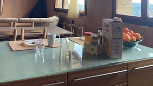 A dining area with a glass table, wooden chairs, a sofa, and a view through large windows. - El Xiprer - El Refugi de la Vall de Biert Total disconnection 25 min from Girona (CANET D'ADRI)