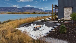 A modern outdoor patio with a fire pit and Adirondack chairs, overlooking a lake and mountains. - Edgewater 103B | 1 Bedroom Suite | Pool | Pineview (Huntsville)