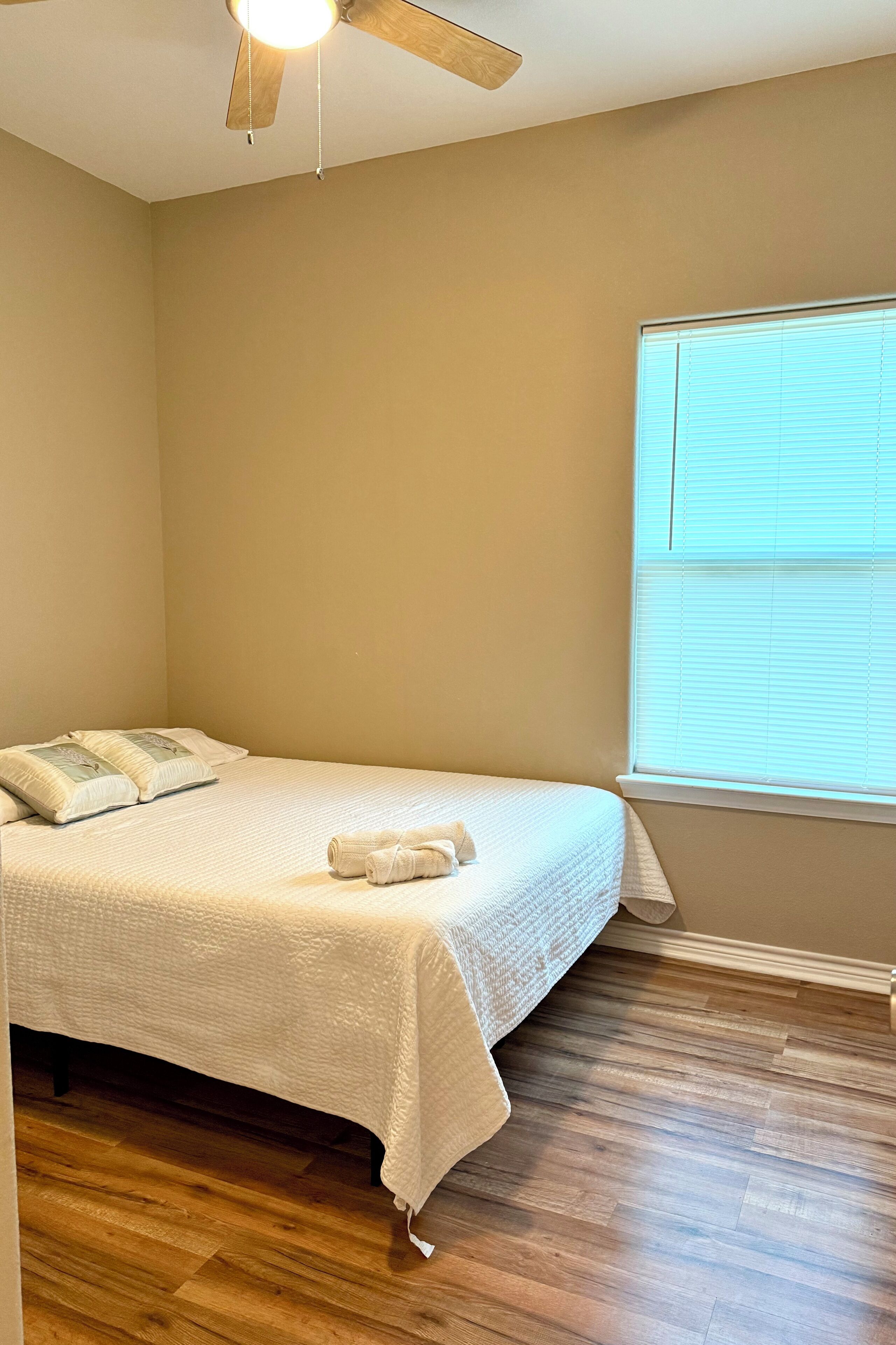 Une chambre à coucher avec un lit, un ventilateur de plafond, une fenêtre avec des stores et un plancher en bois.
