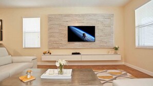 Una sala de estar con una televisión de pantalla plana montada en una pared de madera, un sofá, una mesa de centro con un jarrón de flores y una alfombra estampada.