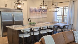 A modern kitchen with a central island, bar stools, a stainless steel refrigerator, pendant lighting, and a dining area with a table and chairs. - Lake Days & Nashville Nights — Private Dock, Game Room & Fire Pit - Room for 12 (Gallatin)