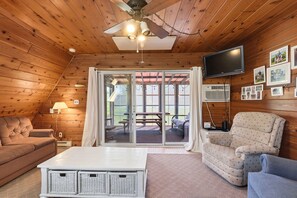 Ein gemütliches Zimmer mit Deckenventilator, einem Flachbildfernseher, einer Couch, einem Sessel, einem Tisch und einer Glastür zu einem Außenbereich.