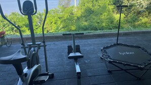 Outdoor exercise equipment, including an elliptical, a stationary bike, and a trampoline, with a view of the sea and greenery in the background.