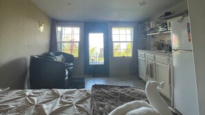 A bedroom with a bed, a chair, a sofa, a kitchen area with cabinets and a refrigerator, and a door leading outside.