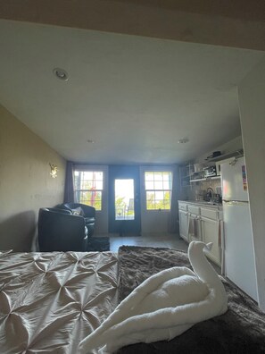 Un dormitorio con cama, una silla, un sofá, una zona de cocina con armarios y un refrigerador, y una puerta que da al exterior.