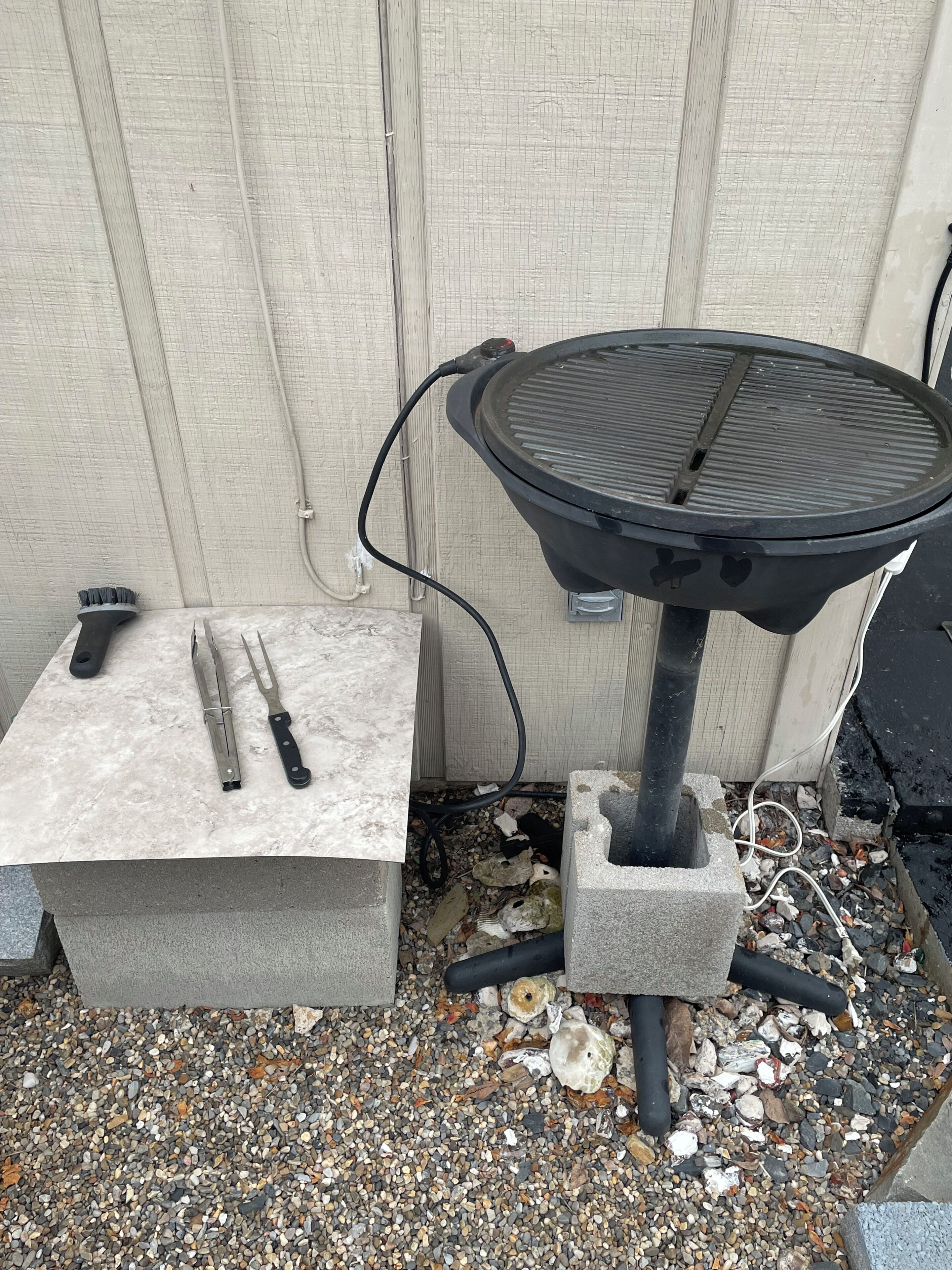 Un barbecue portatif muni d’un cordon d’alimentation, d’une pince et d’un couteau, le tout posé sur un bloc de pierre.
