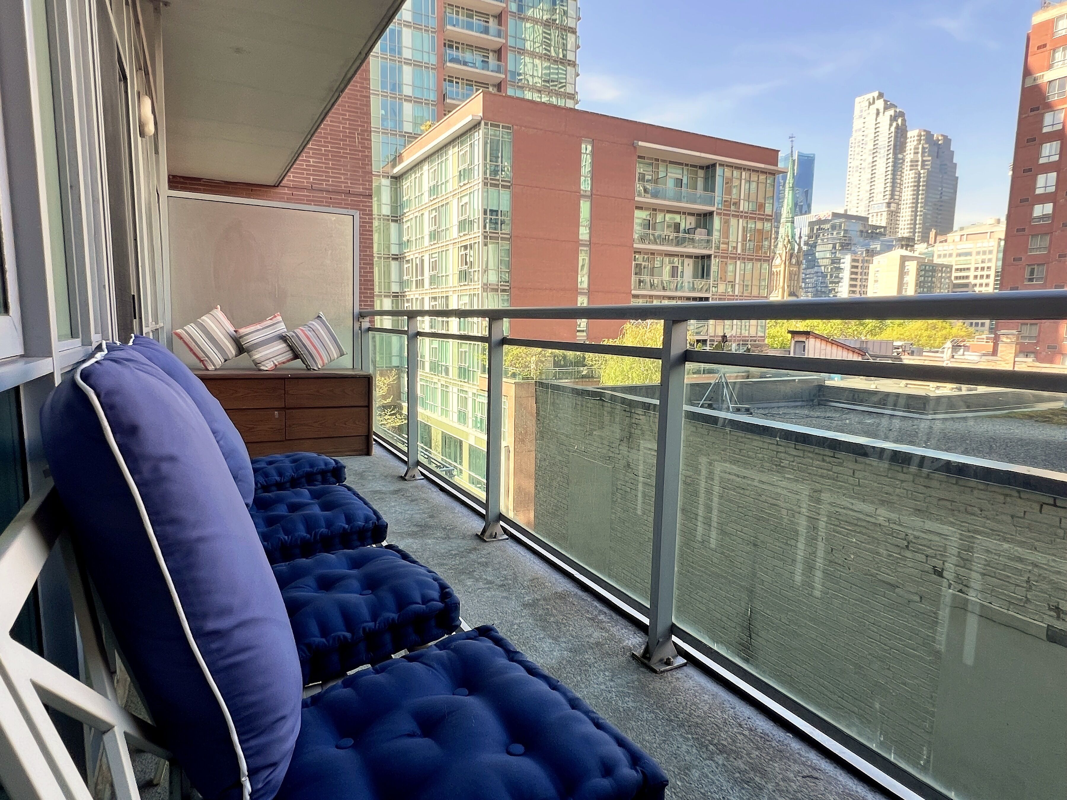 A balcony with a view of a cityscape, a cushioned seating area, and a small wooden table with pillows.