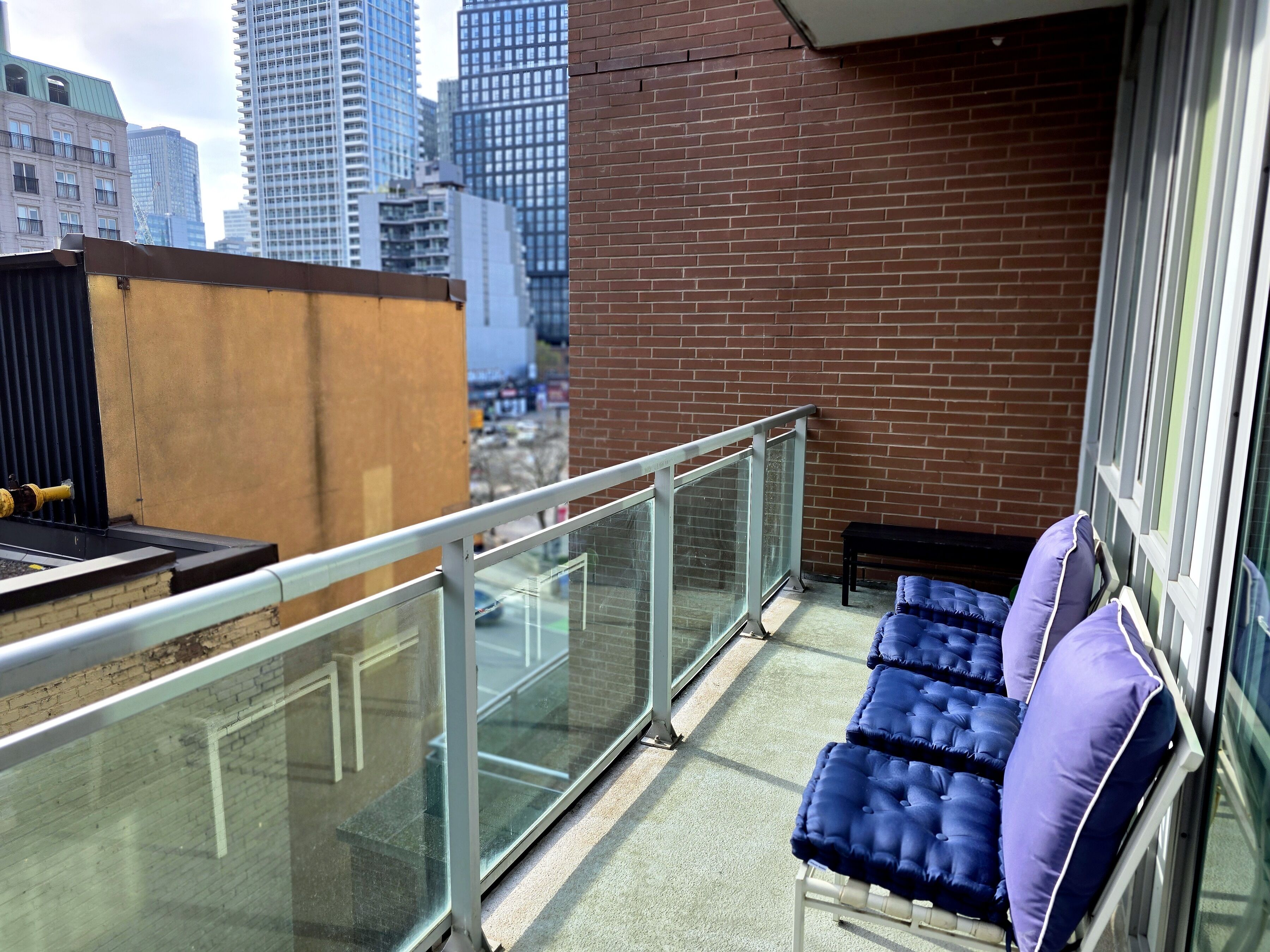 A balcony with a glass railing, a brick wall, and a view of city buildings.