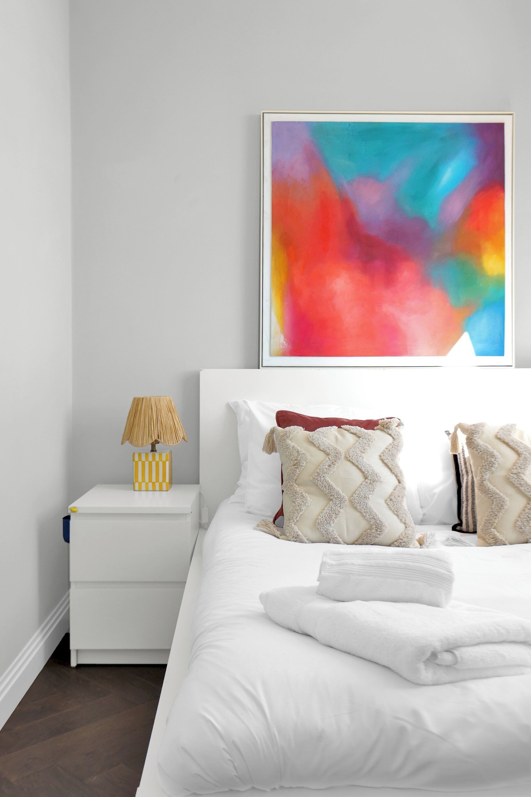 Un dormitorio con una cama blanca, una mesita de noche con una lámpara y un cuadro abstracto colorido en la pared.
