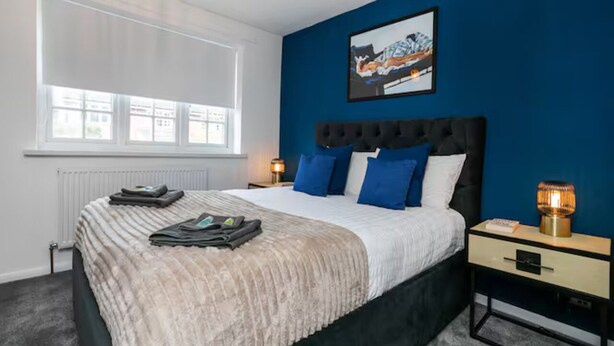 Een slaapkamer met een bed, nachtkastje, lamp, raam en een schilderij aan de muur.