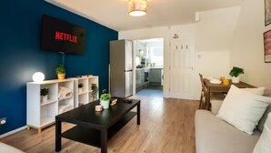 Een woonkamer met een flatscreen televisie, een salontafel, een bank, een boekenkast en een keuken op de achtergrond.