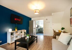 A living room with a flat-screen TV, a coffee table, a sofa, a bookshelf, and a kitchen in the background. - City Centre Home | Fast WiFi | Free Parking - Pass the Keys (Nottingham)