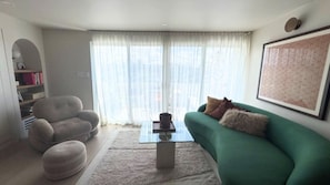 Ein Wohnzimmer mit einem grünen Sofa, einem grauen Sessel, einem Couchtisch und einem großen Fenster mit transparenten Vorhängen.