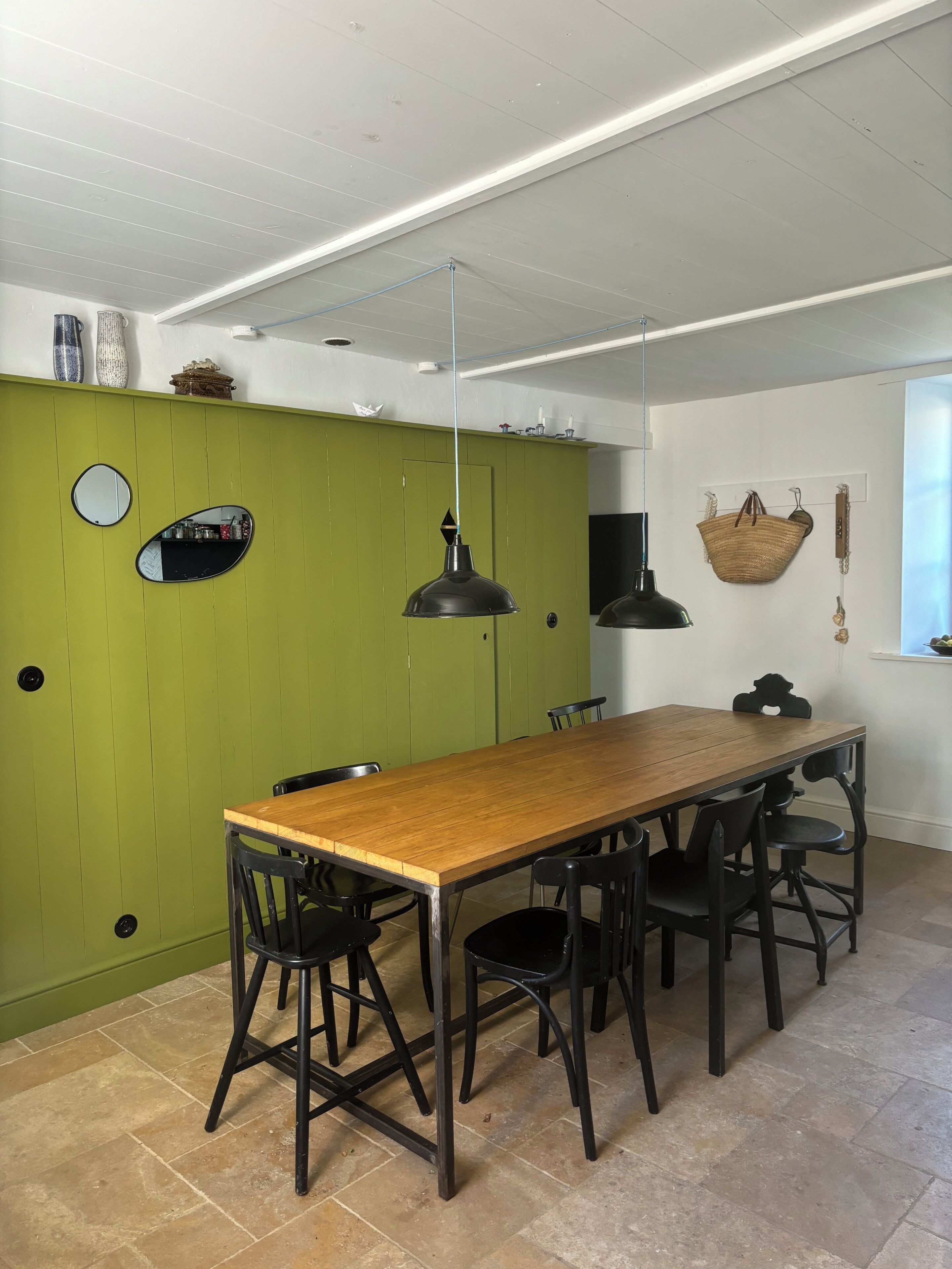Un comedor con mesa de madera y sillas negras, un muro verde decorativo y una cesta colgante.
