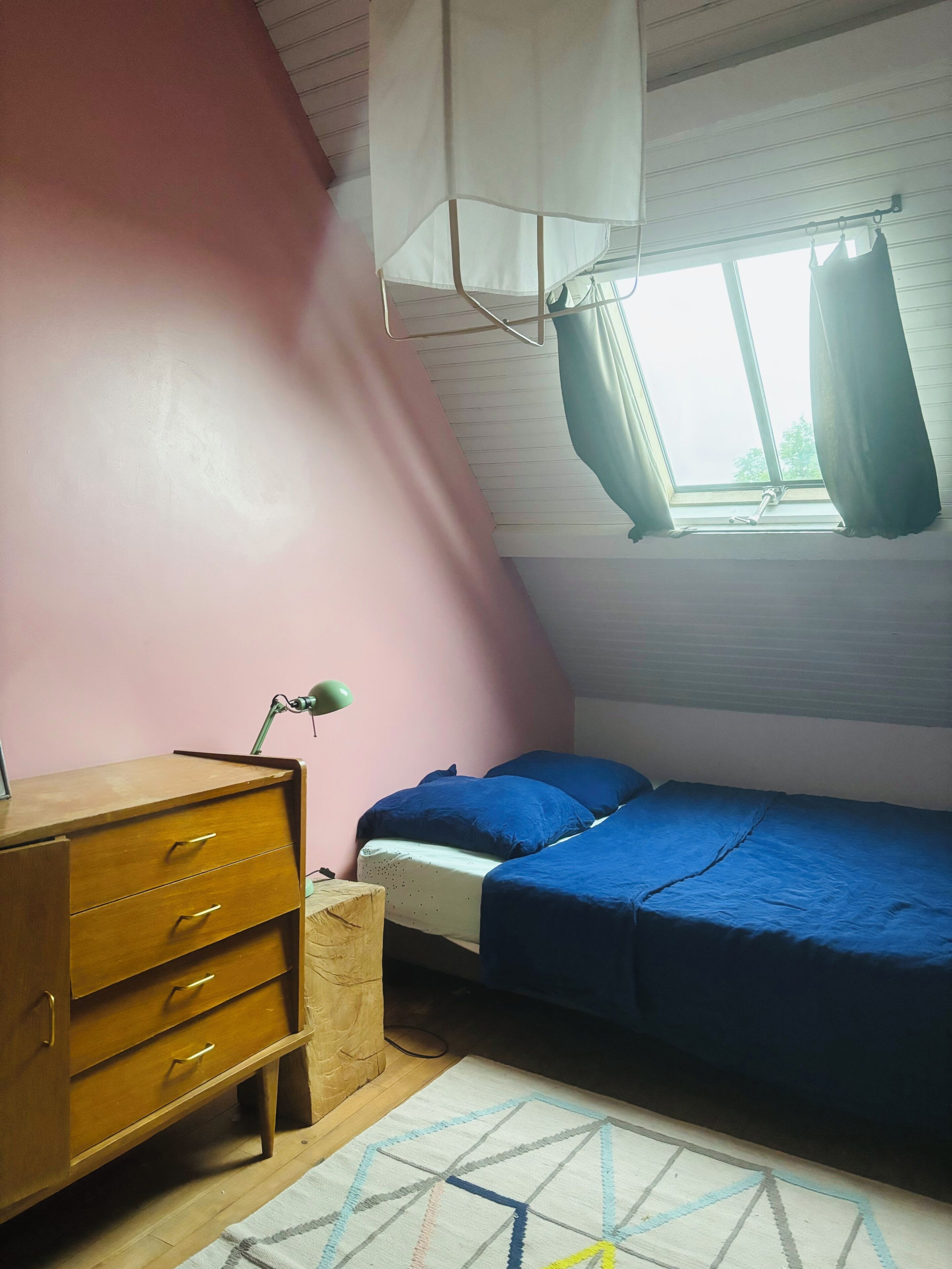 A bedroom with a single bed, a wooden dresser, a desk, and a window with curtains.