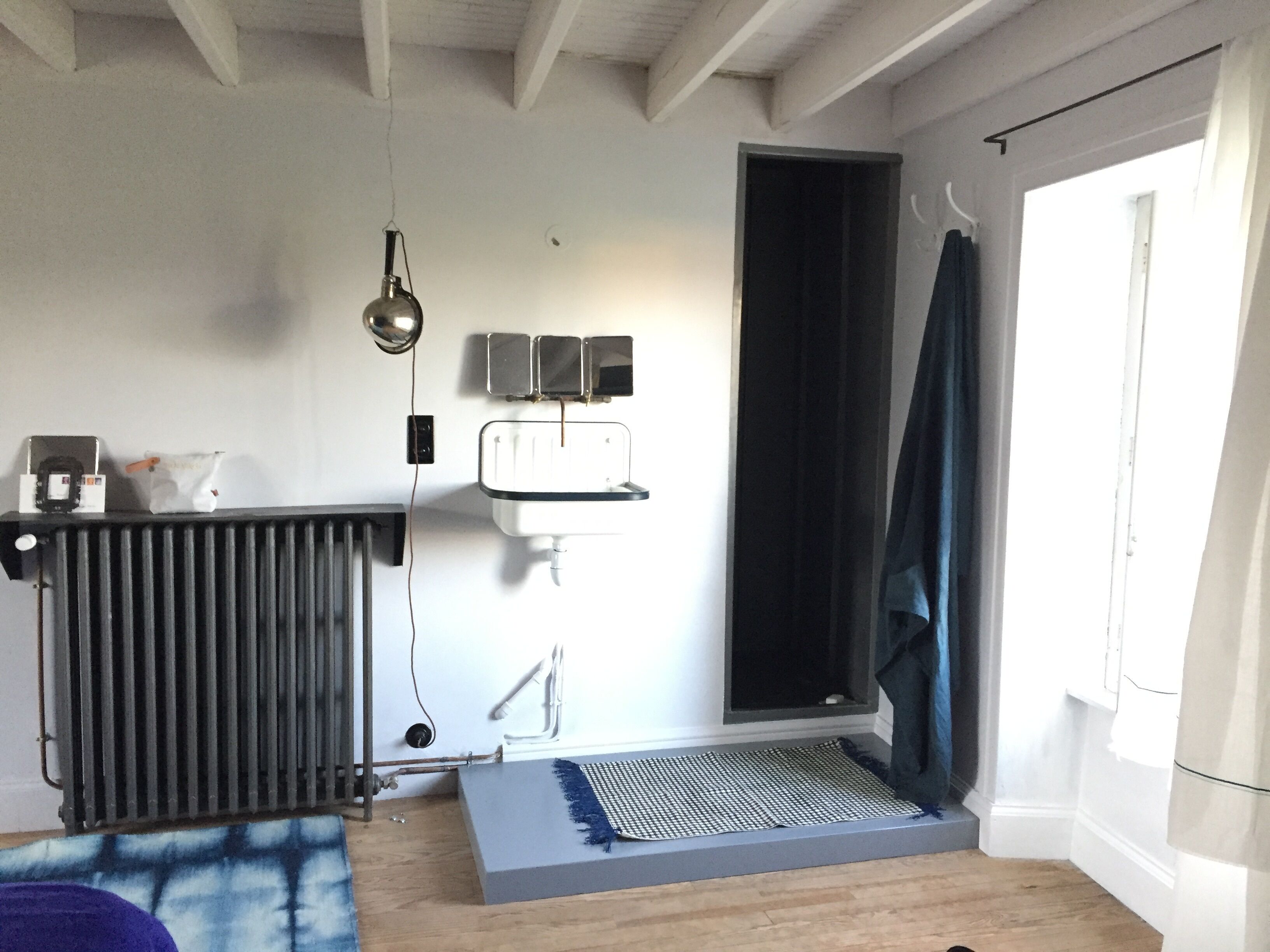 Une pièce avec un radiateur, un lavabo fixé au mur, un luminaire suspendu et une fenêtre avec un rideau.