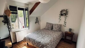 A bedroom with a sloped ceiling, a bed with patterned bedding, a hanging light fixture, a radiator, and a window with a view of greenery. - Charming house (Béthisy-Saint-Martin)