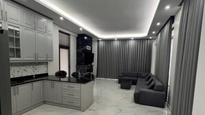 Eine moderne Küche mit Marmorböden, einer integrierten Kochinsel und einem dunkelgrauen Sofa in einem Wohnbereich mit grauen Vorhängen.
