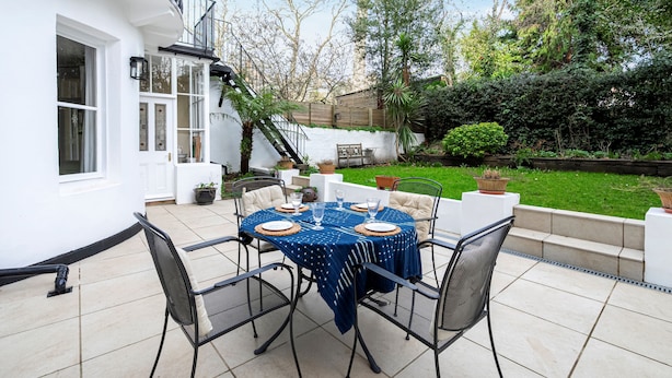 A patio with a round table set for four, surrounded by chairs, and a white building with a balcony in the background. - Elegant 2 bed near Hampstead & Camden (London)