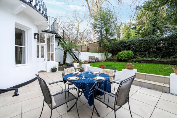 Eine Terrasse mit einem runden Tisch, gedeckt für vier Personen, umgeben von Stühlen, und ein weißes Gebäude mit Balkon im Hintergrund.