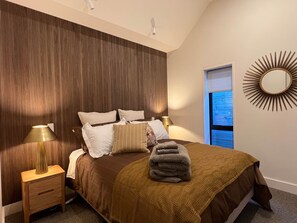 Ein Schlafzimmer mit einem Bett, Nachttischlampen, einem Wandspiegel und einem Fenster mit Jalousien.