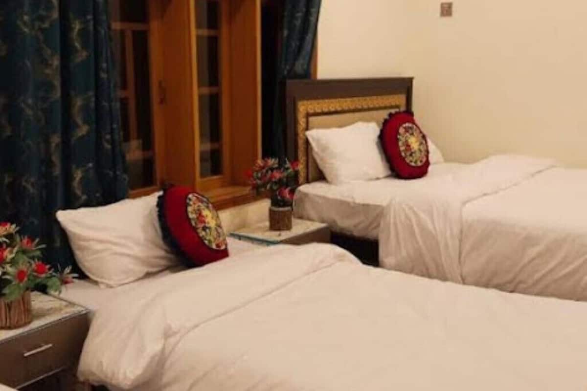 Quarto de hotel com duas camas, uma mesinha de cabeceira, uma janela com cortinas e uma pequena mesa com um vaso de flores.