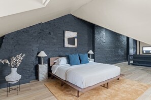 A modern bedroom with a sloped ceiling, a bed with white bedding, blue pillows, and a dark brick wall.