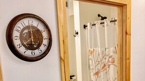 En dörr av trä leder in till ett badrum med en toalett, en duschdraperi med ett blommönster och en väggklocka.