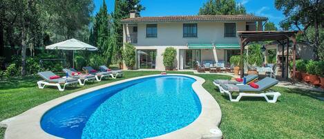A swimming pool with lounge chairs, a covered patio, and a two-story house with a tiled roof.