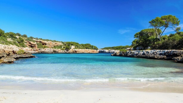 A beach with clear turquoise water, sandy shore, and rocky cliffs with greenery. - Single Storey Villa, A/C, Private Pool - Just a 5min Walk to the Picturesque Cove of Cala Sa Nau! (S´Horta)