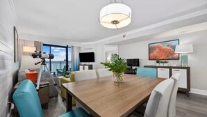 A modern hotel room with a wooden dining table, white and teal chairs, a large window with a view, a flat-screen TV, and a plant on the table. - Park at Your Door! Direct Oceanfront + Waterpark (Myrtle Beach)