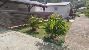 Um edifício com área de estar coberta ao ar livre, um pequeno jardim com plantas de palmeira e um carro estacionado ao fundo.