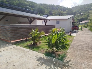 A building with a covered outdoor seating area, a small garden with palm plants, and a parked car in the background. - Bungalow with private Jacuzzi (Deshaies)