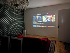Una habitación con una pantalla grande que muestra la interfaz de un servicio de streaming, un sofá rojo, un altavoz pequeño y una pared con estampado.