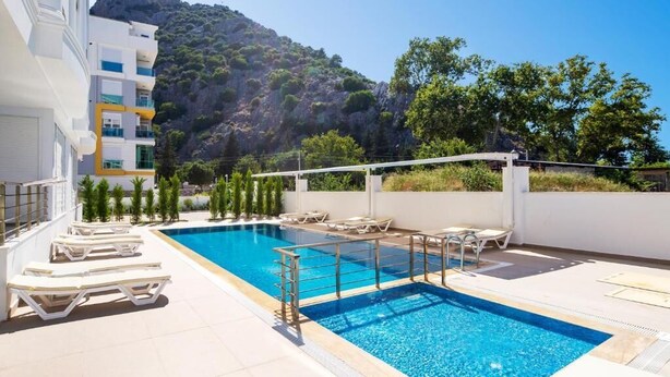 Una piscina con sillas de descanso, un edificio blanco con balcones y un paisaje montañoso de fondo.