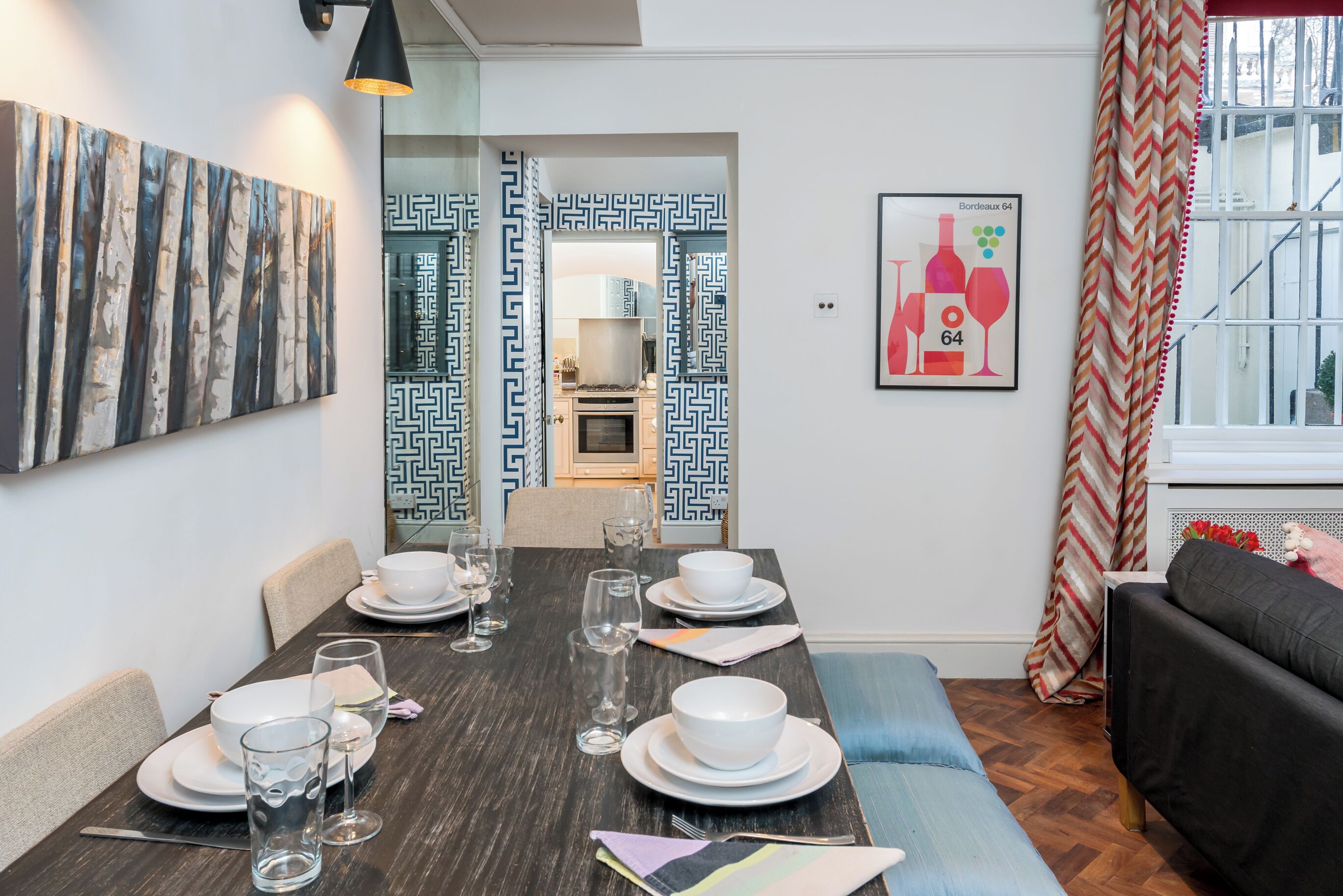 A dining area with a dark wooden table set for four, white plates, and clear glasses. A black sofa and a patterned curtain are visible in the background.