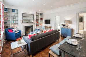A living room with a fireplace, a sofa, a dining table set for two, and a television mounted on the wall. - Charming 1BR Retreat in Pimlico (London)