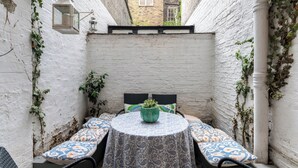 A small outdoor seating area with a round table and chairs, surrounded by white brick walls and covered with a patterned tablecloth. - Charming 1BR Retreat in Pimlico (London)