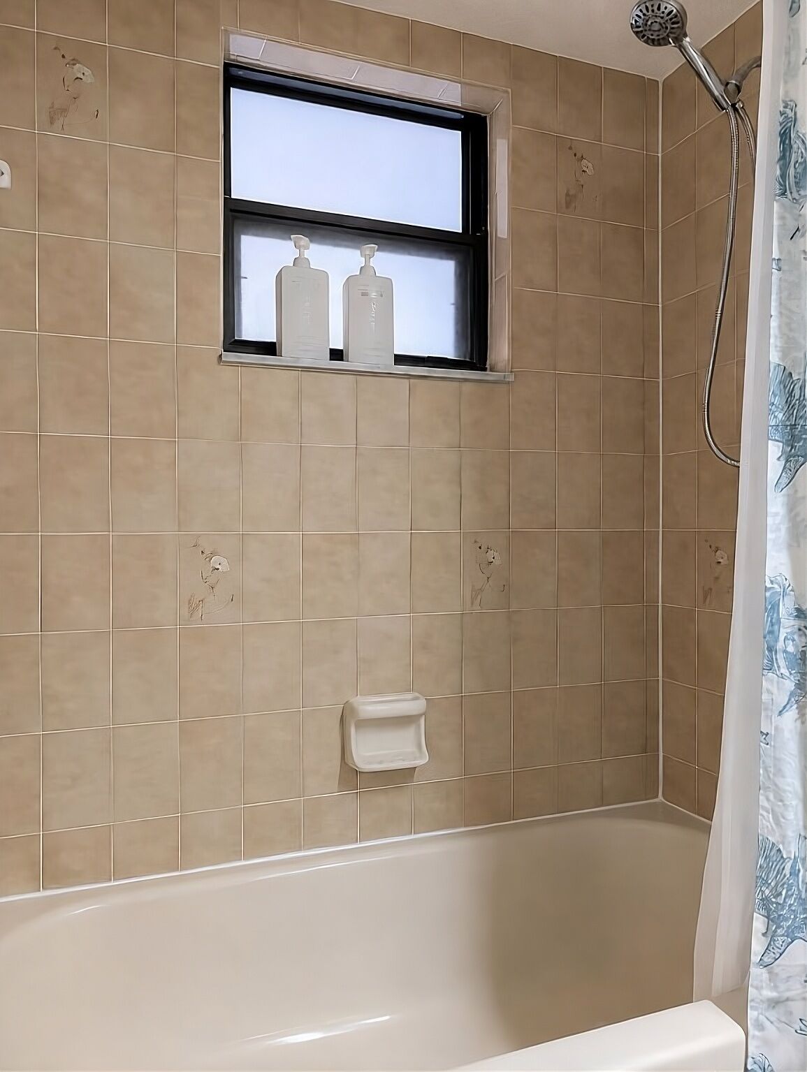 A bathroom with beige tiles, a window with two bottles, a soap dish, and a shower curtain.